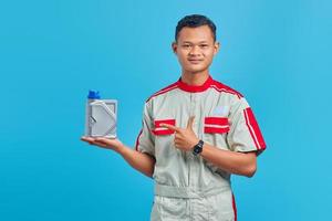 retrato de um jovem mecânico asiático sorridente apontando para uma garrafa de plástico de óleo de motor com o dedo sobre fundo azul foto