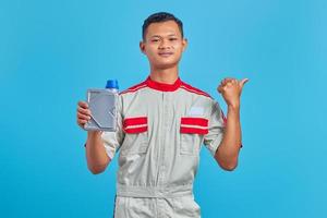 retrato de um jovem mecânico asiático sorridente, mostrando a garrafa de plástico de óleo de motor e apontando para o espaço vazio sobre um fundo azul foto