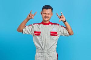 retrato de um jovem mecânico asiático gritando com uma expressão maluca fazendo o símbolo do rock com as mãos sobre o fundo azul foto