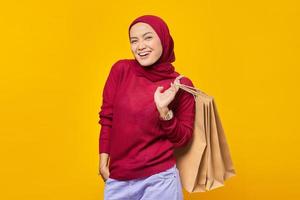 Mulher asiática jovem feliz e confiante sorrindo para a câmera com as mãos nos bolsos e segurando sacolas de compras no fundo amarelo foto