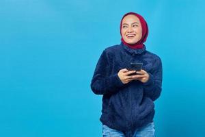 retrato de uma jovem sorridente asiática segurando um telefone celular e olhando para o lado sobre fundo azul