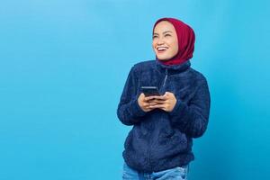 Retrato de uma jovem asiática sorridente, segurando um telefone celular e olhando para o lado sobre fundo azul.