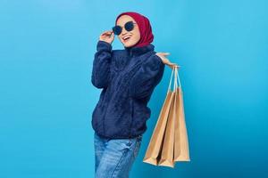 alegre jovem asiática usando óculos escuros e segurando sacolas de compras sobre fundo azul foto