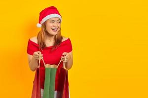 alegre jovem asiática com chapéu de Papai Noel abrindo a sacola de compras e olhando para o fundo amarelo