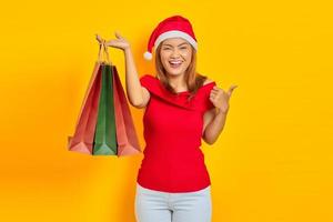 retrato de alegre jovem asiática com chapéu de Papai Noel mostrando sacolas de compras e gesto de aprovação sobre fundo amarelo