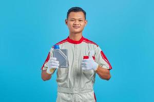 retrato de um jovem mecânico asiático sorridente, mostrando a garrafa de plástico de óleo de motor com o polegar para cima gesto isolado sobre fundo azul foto