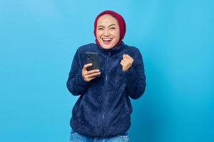 Mulher asiática jovem feliz usando telefone celular com gesto de mão bem-sucedido em fundo azul foto