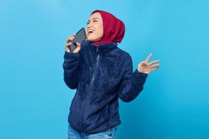 Mulher asiática jovem e feliz curtindo cantar usando o smartphone sobre fundo azul foto
