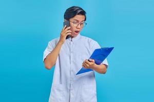 retrato de enfermeiro surpreso falando no smartphone enquanto segura a prancheta isolada sobre fundo azul foto