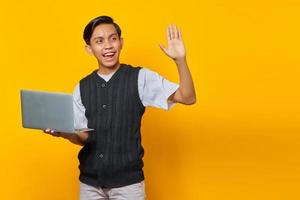 retrato de um jovem asiático sorridente segurando um laptop e gesticulando com a mão da onda sobre fundo amarelo