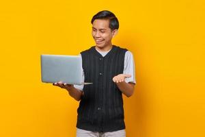 feliz bonito jovem empresário olhando para o laptop e levantando a mão sobre fundo amarelo foto