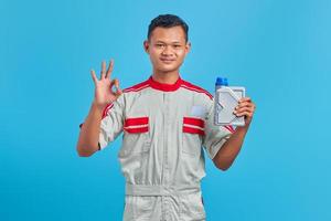retrato de um jovem mecânico asiático sorridente segurando uma garrafa de plástico de óleo de motor e mostrando aprovação com o polegar para cima sobre o fundo azul foto