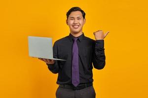 empresário asiático sorridente segurando laptop enquanto aponta o polegar para o espaço da cópia em fundo amarelo foto