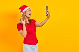 alegre jovem asiática usando chapéu de natal falando uma selfie com o celular em fundo amarelo