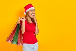 animada jovem asiática com chapéu de Papai Noel segurando sacolas de compras e olhando para o espaço da cópia isolado sobre fundo amarelo foto