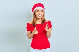 retrato de uma jovem asiática sorridente com vestido vermelho e chapéu de Natal apontando o dedo para a câmera, isolado no fundo branco