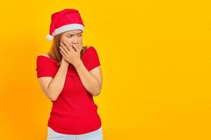 jovem asiática surpresa com chapéu de Natal cobrindo a boca com as mãos em fundo amarelo foto