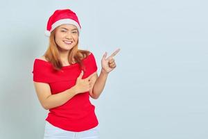 retrato de uma jovem asiática sorridente com chapéu de Natal apontando o dedo para o espaço da cópia sobre fundo branco