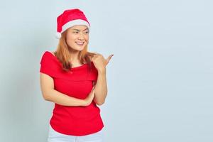 foto de uma linda garota asiática com chapéu de Natal com expressão facial sorridente e apontando para o lado no fundo branco