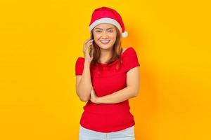 Mulher asiática jovem sorridente com chapéu de Natal falando no smartphone e olhando para a câmera sobre fundo amarelo foto