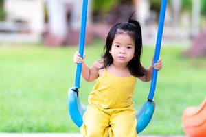 menina asiática brincar no balanço azul. criança com vestido amarelo. criança de 3 anos se diverte no playground. foto