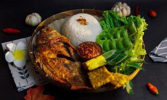 um pacote de arroz, peixe frito e alguns vegetais frescos em um fundo preto