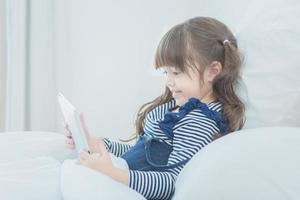 menina bonitinha gosta de assistir desenhos animados em um tablet inteligente com uma boneca bonita enquanto está sentada na cama, no quarto das crianças em casa foto