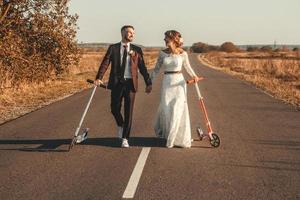 casal de noivos sorridente andando de scooters ao longo da estrada fora da cidade ao pôr do sol. lugar para texto ou publicidade foto