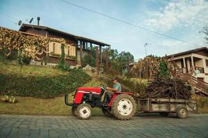 bento goncalves, brasil - 13 de julho de 2019. trator puxando carreta com mato em uma estrada asfaltada de pedra, próximo a uma casa de madeira perto de bento goncalves. uma simpática cidade do interior famosa por sua produção de vinho. foto