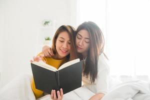 belas jovens mulheres asiáticas lgbt lésbicas casal feliz sentado na cama lendo livro juntos perto da janela no quarto em casa. conceito de casal de lésbicas lgbt juntos dentro de casa. passar um bom tempo em casa. foto
