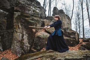 jovem de quimono preto praticando artes marciais com uma espada nas rochas e no fundo da floresta foto