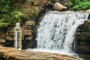 garrafa de plástico transparente com água fica em um tronco de madeira contra o fundo do rio e da cachoeira