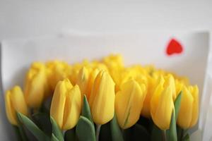 lindo grande buquê de tulipas amarelas em um pacote branco e confete de coração vermelho na mesa branca close-up. parabéns. fundo festivo. copie o espaço.