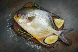 Peixe pomfret fresco com ervas, especiarias, alecrim e limão na tábua de madeira e fundo de placa preta - peixe pomfret preto cru foto