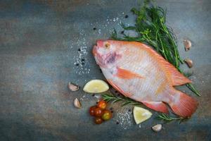 peixe fresco com ervas especiarias alecrim e tomate com alho e limão - tilápia vermelha de peixe cru no fundo da placa escura foto