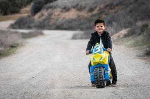 um garotinho feliz dirigindo uma motocicleta de brinquedo, vestido com uma jaqueta de couro de motociclista em uma estrada rural foto