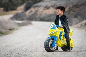 um garotinho feliz dirigindo uma motocicleta de brinquedo, vestido com uma jaqueta de couro de motociclista em uma estrada rural foto