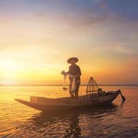 silhueta de pescador asiático segurando uma rede para pegar peixes de água doce no lago natural durante o nascer do sol foto