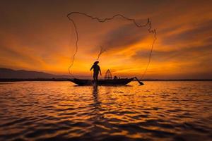 pescador asiático em um barco de madeira lançando uma rede para pegar peixes de água doce no rio da natureza no início da manhã, antes do nascer do sol