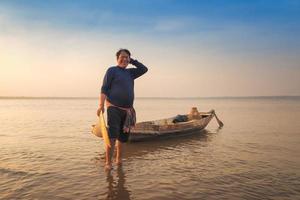 pescador asiático sorrindo com um barco de madeira no rio da natureza no início da manhã antes do nascer do sol foto