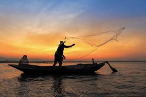 silhueta do pescador asiático em um barco de madeira em ação, jogando uma rede para a captura de peixes de água doce no rio da natureza no início da manhã, antes do nascer do sol foto