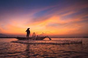 pescador asiático em um barco de madeira, jogando uma rede para pegar peixes de água doce no rio da natureza, de manhã cedo, antes do nascer do sol foto