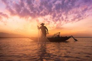 silhueta de pescador asiático segurando uma rede para pegar peixes de água doce no lago natural durante o nascer do sol foto