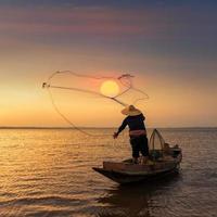 pescador asiático em um barco de madeira jogando uma rede para pegar peixes de água doce no rio da natureza no início durante o nascer do sol