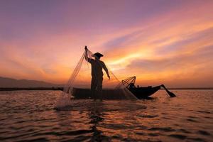 pescador asiático em um barco de madeira jogando uma rede para pegar peixes de água doce no rio da natureza no início durante o nascer do sol