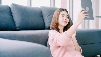 mulher asiática usando tablet enquanto estava deitada no sofá em sua sala de estar. feliz feminino usar tablet para videochamada com os amigos ou namorado em casa. foto