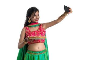 linda jovem feliz tirando uma selfie com lâmpada de argila ou diya durante o festival de diwali luz usando um smartphone em um fundo branco foto