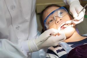 dentista usando utensílios odontológicos para limpar os dentes de uma criança asiática e tratar cáries na clínica com um assistente em pé atrás do paciente. conceito de odontologia e saúde foto