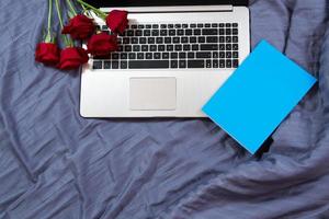 laptop com vista de cima, bloco de notas azul vazio, buquê de flores vermelho no fundo do espaço da cópia da cama foto