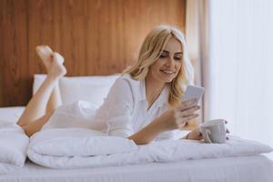 Mulher feliz usando um telefone celular enquanto está deitada na cama e bebe café da manhã foto
